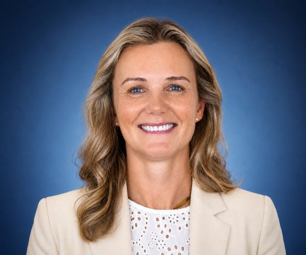 Professional headshot of a smiling blonde woman in a beige blazer against a blue gradient background, suitable for corporate use.